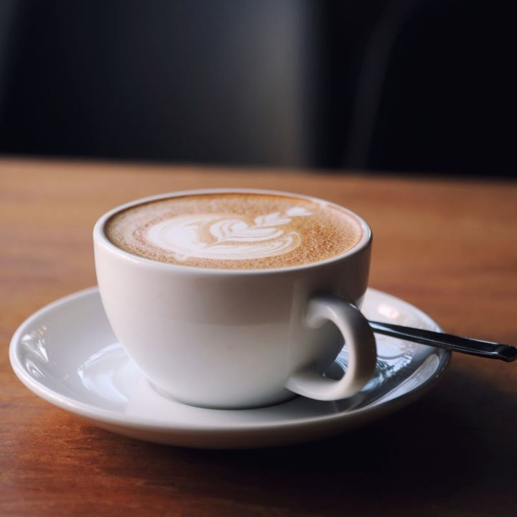 A delicious cup of Cafe Latte served in a white cup with elegant latte art on top, placed on a saucer with a spoon on a wooden table.