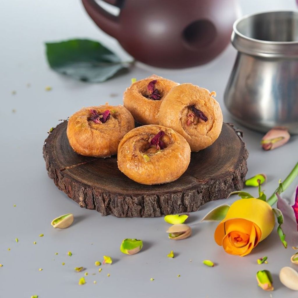 Four pieces of traditional Indian sweet delicacy garnished with rose petals and pistachios, placed on a rustic wooden slab with scattered nuts and a yellow rose in the background.