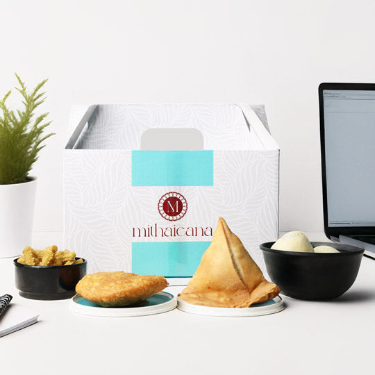 mithaicana indian snack gift box with samosas, pakoras, and laddoos, on a white table with a laptop in the background