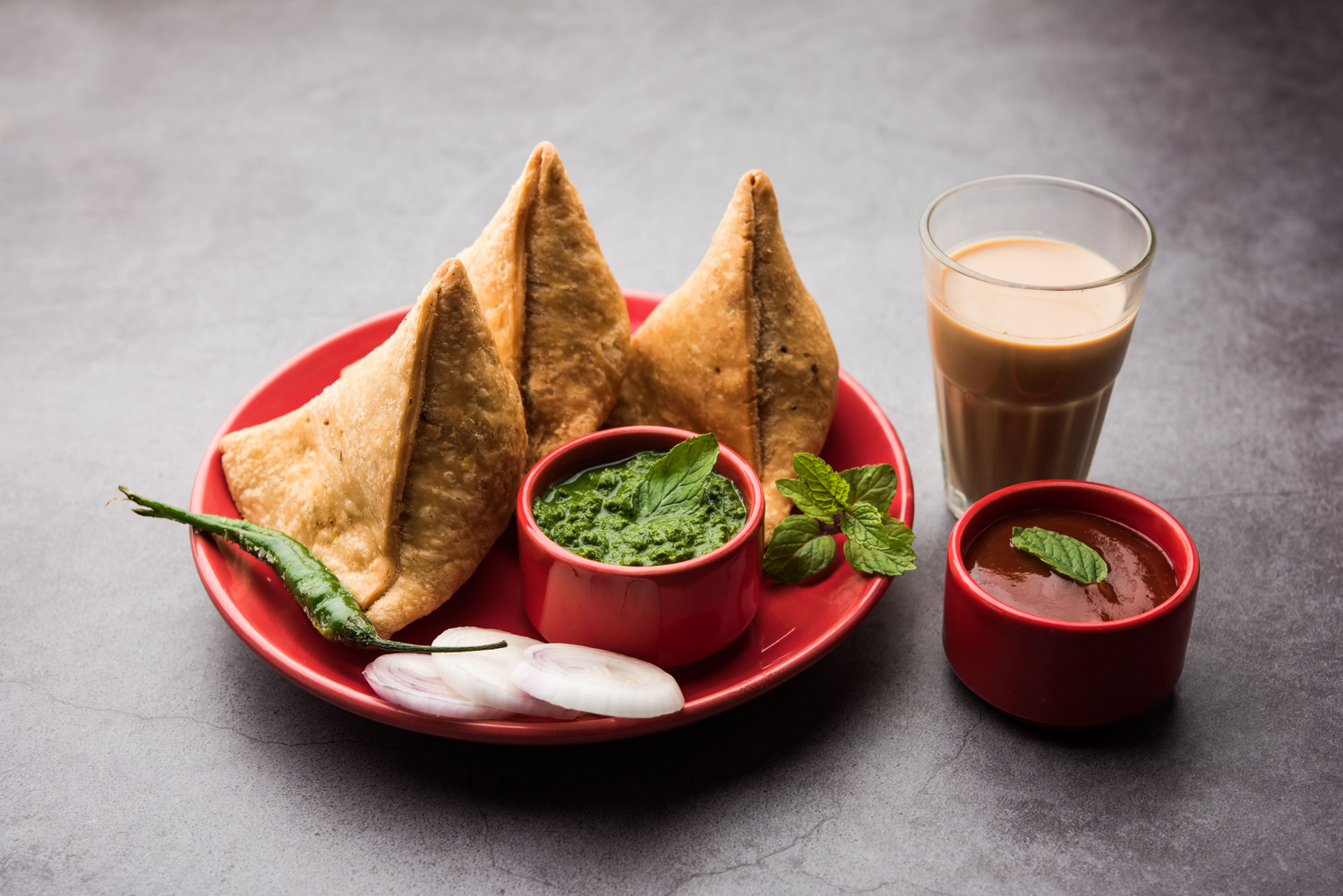 samosas served with onion rings and green chilies, with green and red chutney as sides